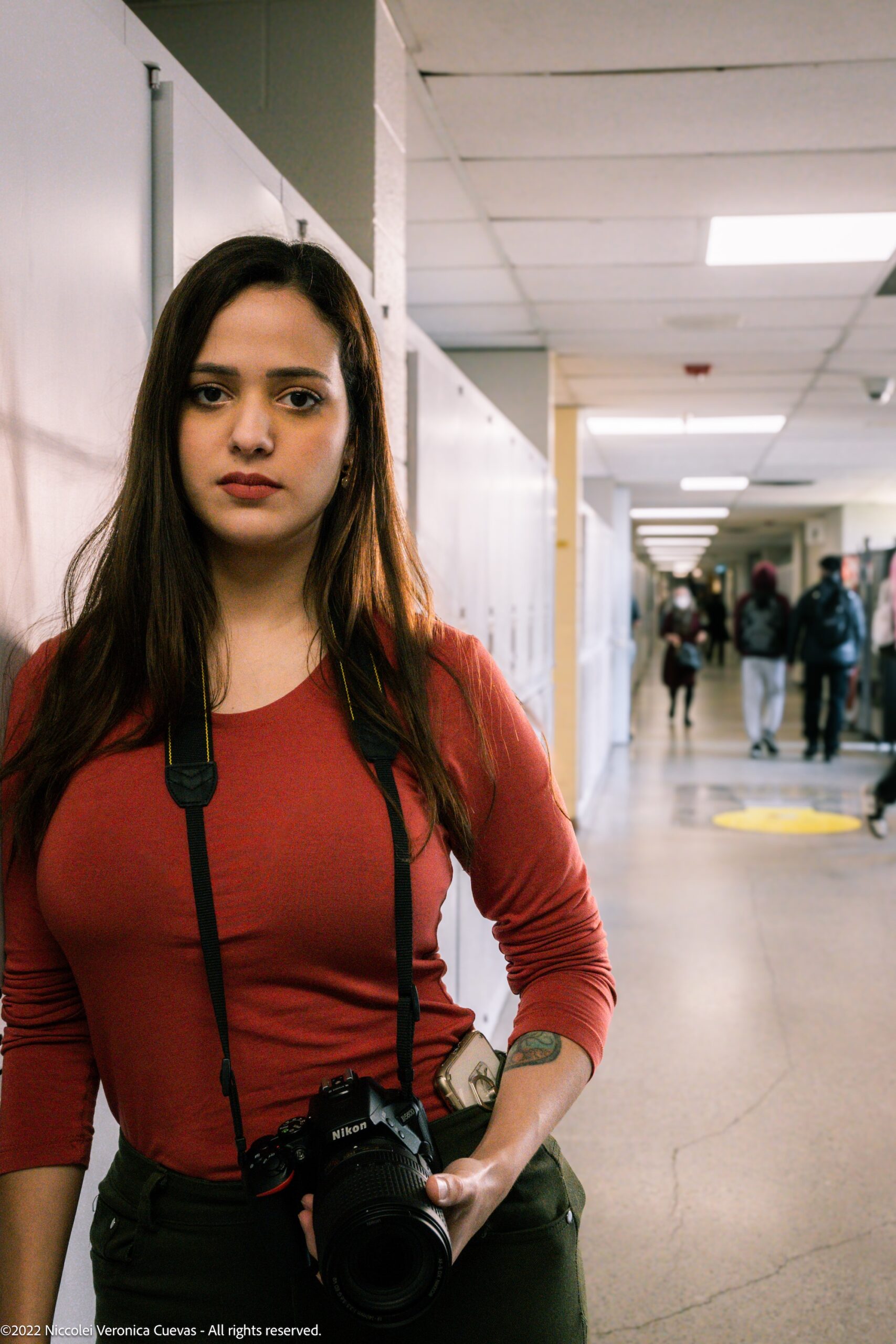 beautiful lady wearing an orange shirt leaning on school locker while holding a dslr, staring at camera.