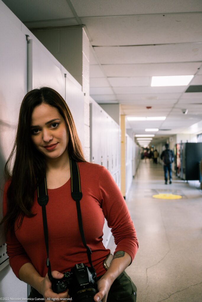 beautiful lady wearing an orange shirt leaning on school locker while holding a dslr, smiling at camera.