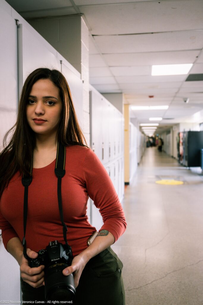beautiful lady wearing an orange shirt leaning on school locker while holding a dslr, smiling at camera.